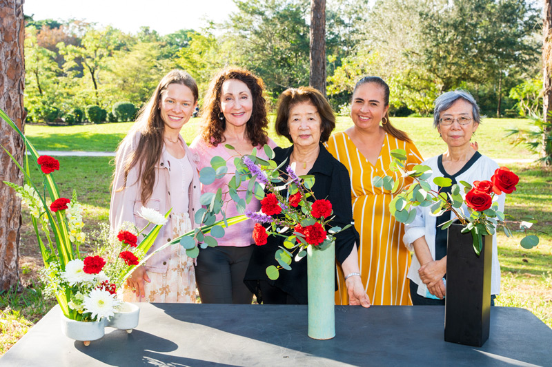 A flower arranging teacher stands proudly with her students, showing off their beautifully arranged floral creations. 