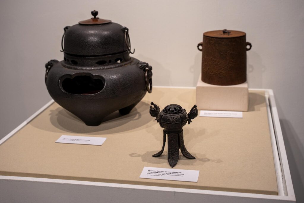Front Object: Iron-cast incense burner in the shape of a chrysanthemum with four horses; back left object: large iron brazier used to heat water during tea ceremonies; back right object: iron fire bucket with design featuring clouds and rain